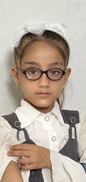 Portrait von Shaha, einem kleinen Mädchen. Sie ist schick angezogen und streckt ihren verletzten Arm nach vorne. Sie schaut dabei ernst in die Kamera.. 